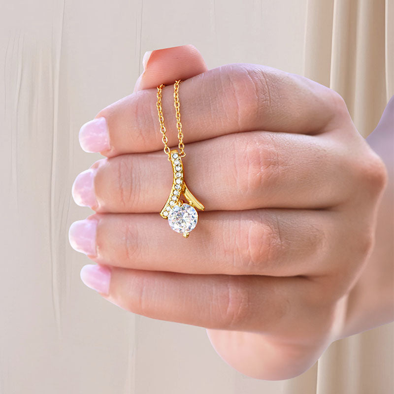 Gold necklace with a ribbon-shaped pendant padded with cubic Zirconia stones and one big round cut Zirconia stone centered, held by a hand against a neutral background