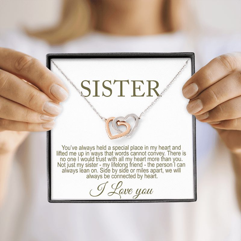 Necklace with interlocking heart pendants held by hands on a card with text for a sister.