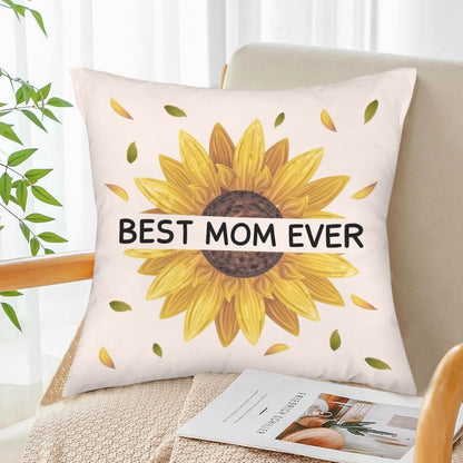 A square off-white throw pillow with a large yellow sunflower illustration and bold text reading "BEST MOM EVER" across the center, styled on a wooden armchair next to a green houseplant and a open magazine.   
