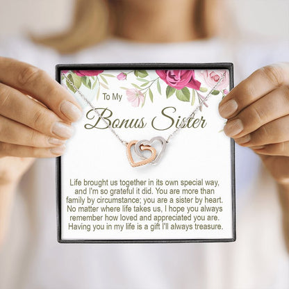 A person holding a gift box containing a To My Bonus Sister interlocking rose gold and silver heart necklace with a floral message card.