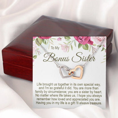 A closed mahogany-style wooden gift box with a "To My Bonus Sister" floral message card and interlocking heart necklace displayed on top against a soft white draped background.   
