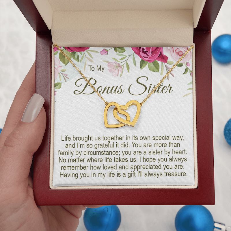An open mahogany-style wooden gift box  held by a hand, containing a gold interlocking heart necklace with a "To My Bonus Sister" floral message card, accented with blue ornaments.   
