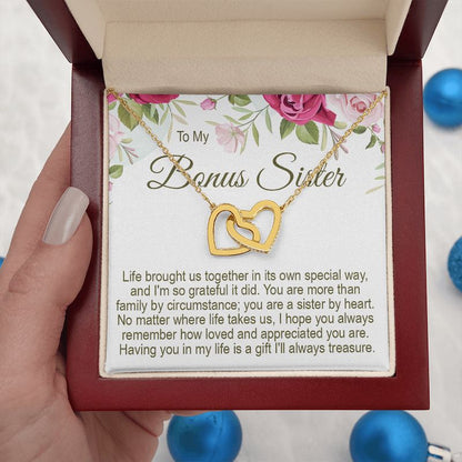 An open mahogany-style wooden gift box  held by a hand, containing a gold interlocking heart necklace with a "To My Bonus Sister" floral message card, accented with blue ornaments.   