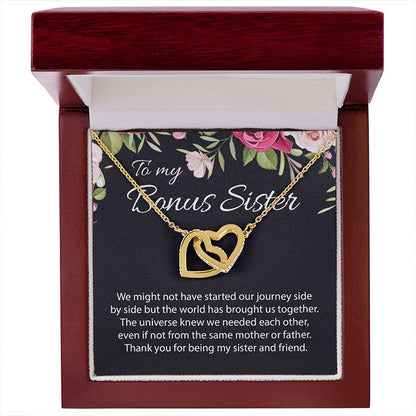 A gold double-heart pendant necklace presented inside an open reddish-brown gift box. The necklace is displayed on a floral message card that reads 'To my Bonus Sister' with a sentimental message below, set against a plain white background.