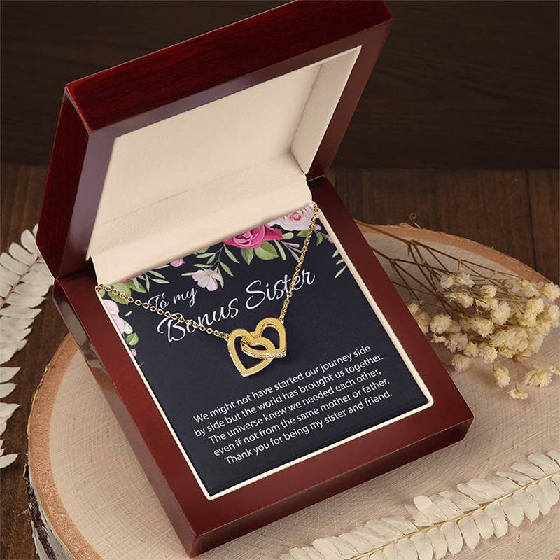An open dark reddish-brown wooden gift box displaying a two gold heart necklace on a floral printed message card. The card reads 'To my Bonus Sister' and includes a sentimental message. The gift box is presented on a natural wooden surface.