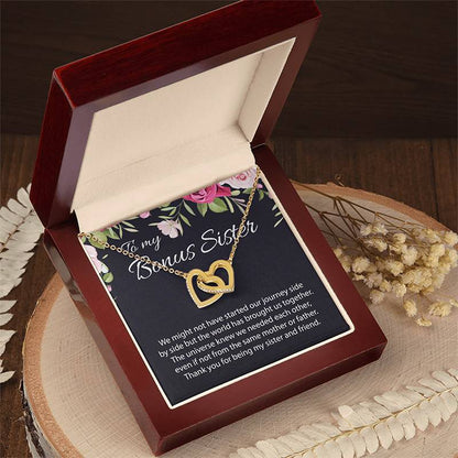 An open dark reddish-brown wooden gift box displaying a two gold heart necklace on a floral printed message card. The card reads 'To my Bonus Sister' and includes a sentimental message. The gift box is presented on a natural wooden surface.