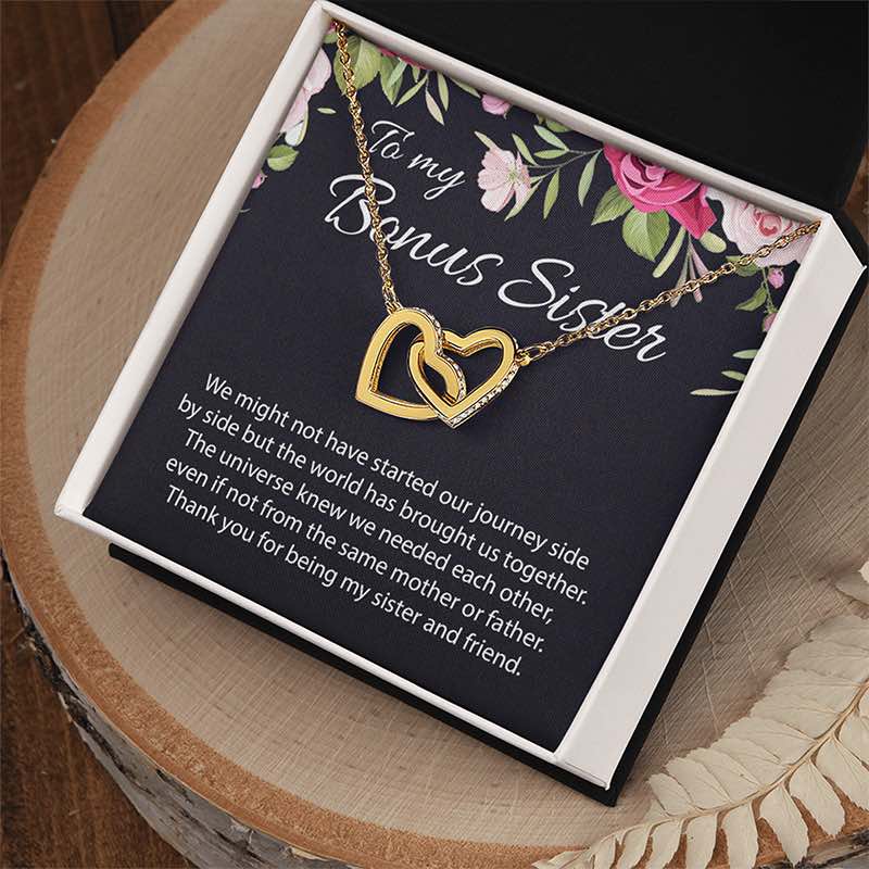 A gold double-heart pendant necklace displayed inside an open gift box with a dark floral message card. The card reads 'To my Bonus Sister' and includes a sentimental message.