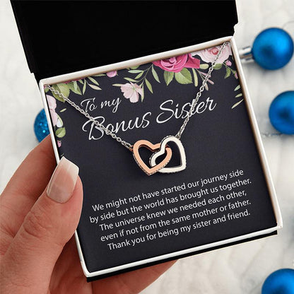 A person's hand holds an open gift box containing a necklace with two interlocking heart pendants, one rose gold and one silver, displayed on a floral message card addressed 'To my Bonus Sister'.