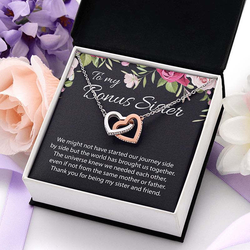 A gift box containing a double-heart pendant necklace with one rose-gold heart and one silver heart, displayed on a floral message card that reads 'To my Bonus Sister' with a sentimental message. The box is styled with pink and purple flowers on a white surface.