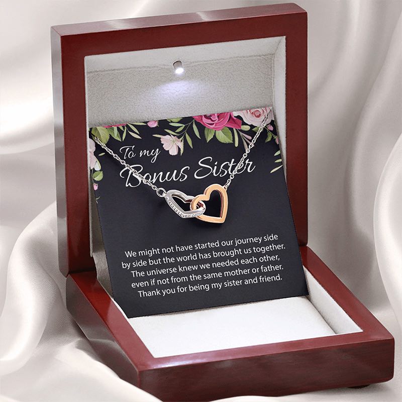 A personalized heart necklace with message card gift in a wooden box against a white satin lining. The necklace features two interlocking hearts, one rose gold and one silver, displayed on a black floral card with a sentimental message for a 'Bonus Sister'.