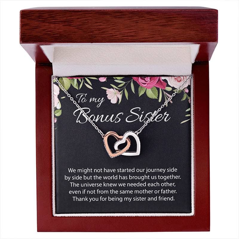 A heart-shaped double pendant necklace in rose gold and silver, displayed on a floral card inside an open reddish-brown wooden gift box. The card reads 'To my Bonus Sister' and includes a sentimental message.
