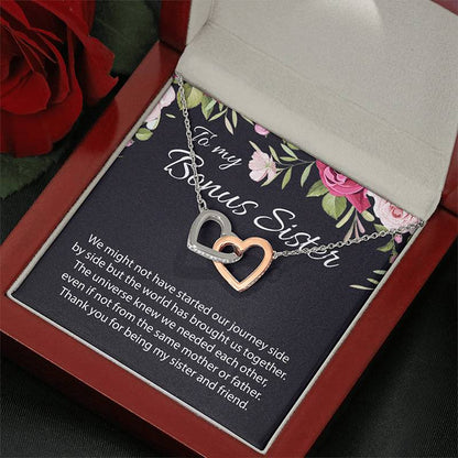 A double heart pendant necklace with one silver heart and one rose gold heart, displayed on a floral message card inside an open red gift box. The card reads 'To my Bonus Sister' and is styled with a red rose in the corner.