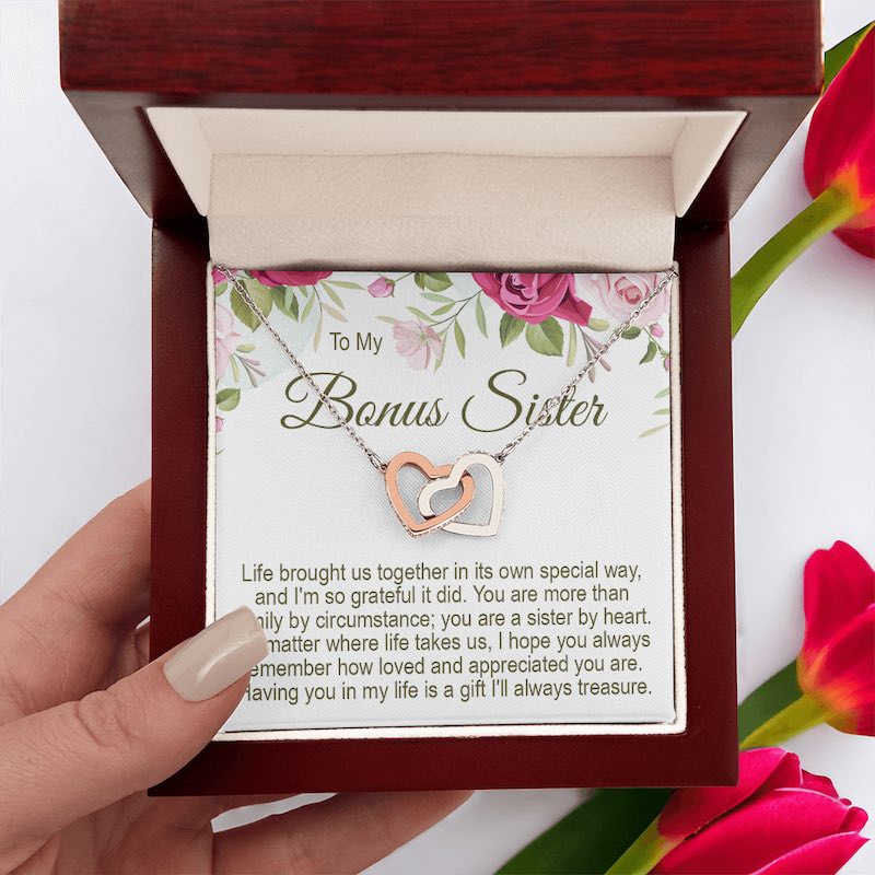 An open mahogany-style wooden gift box, held by a hand, containing a To My Bonus Sister interlocking rose gold and silver heart necklace with a floral message card, next to pink tulips.