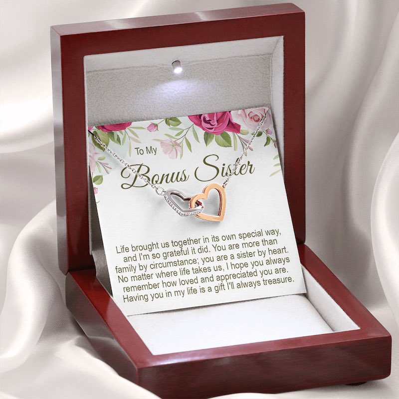 An illuminated luxury mahogany-style wooden gift box holding a silver and rose-gold interlocking heart necklace, displayed against a soft white draped background with a floral message card reading "To My Bonus Sister".   