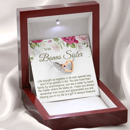 An illuminated luxury mahogany-style wooden gift box holding a silver and rose-gold interlocking heart necklace, displayed against a soft white draped background with a floral message card reading "To My Bonus Sister".   