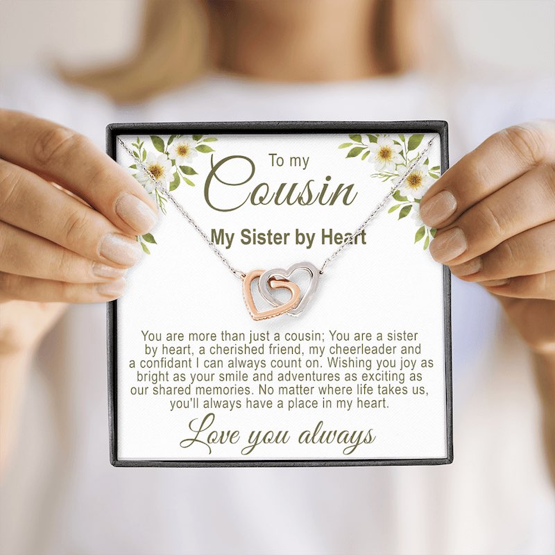 A square black gift box featuring an interlocking rose gold and silver hearts necklace with a floral cousin message card, held by a person in a blurry background.