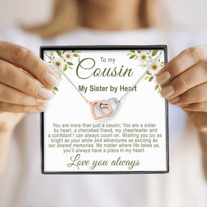 A square black gift box featuring an interlocking rose gold and silver hearts necklace with a floral cousin message card, held by a person in a blurry background.