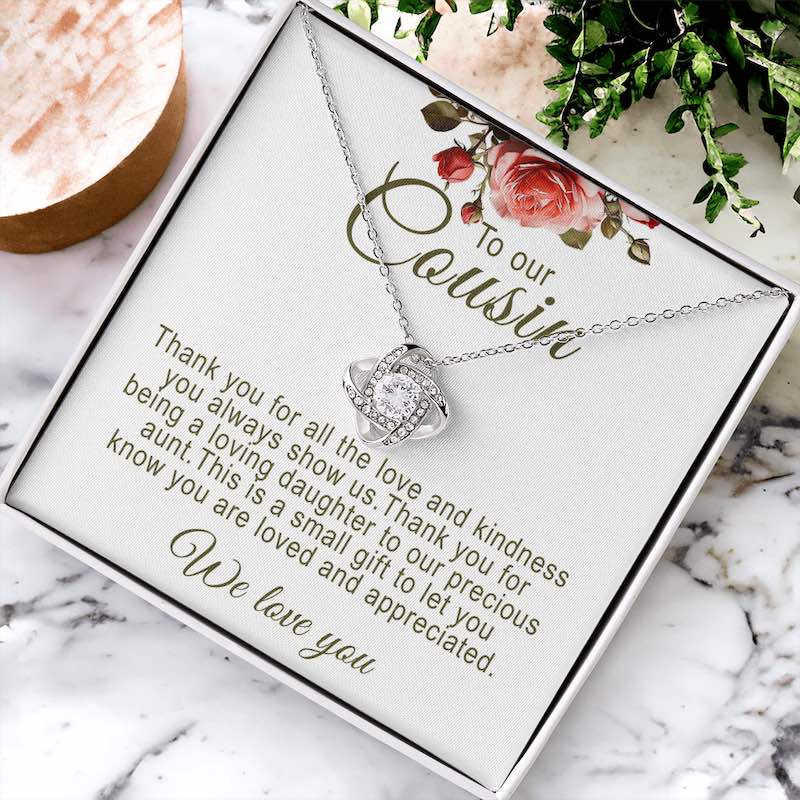Silver love knot pendant necklace in a white gift box with floral artwork and message card, displayed flat on a marble surface next to green foliage and decorative accents.