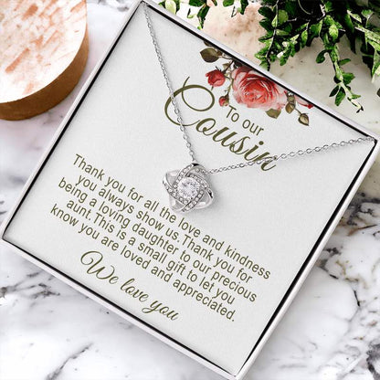 Silver love knot pendant necklace in a white gift box with floral artwork and message card, displayed flat on a marble surface next to green foliage and decorative accents.