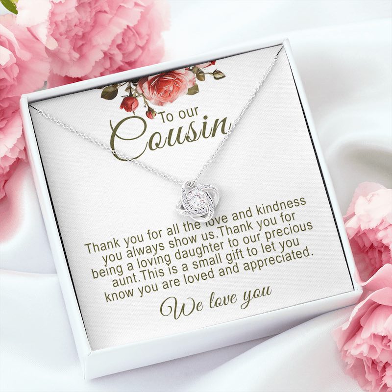 A silver love knot pendant necklace featuring a central round cubic zirconia surrounded by pavé crystal accents. The necklace sits on a white message card in an open white gift box, displayed on a soft white silk background surrounded by fresh pink carnation blossoms. The card features red rose artwork at the top and reads: "To our Cousin.