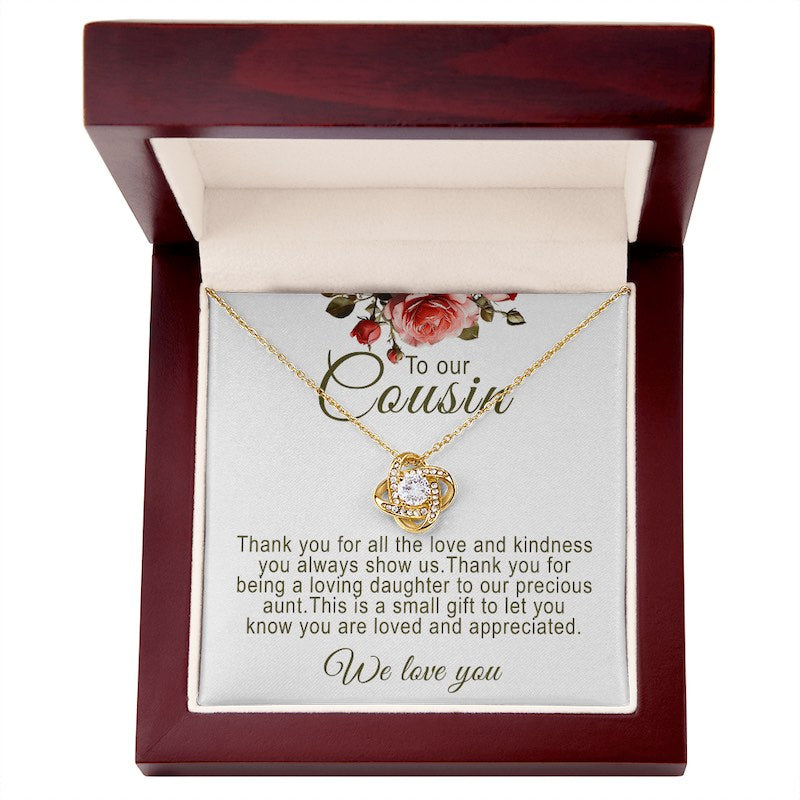 A gold love knot pendant necklace featuring a central round cubic zirconia surrounded by pavé accent crystals. The necklace is presented on a white message card for cousin, inside an open mahogany-finish luxury wooden gift box, accented with red rose artwork at the top.