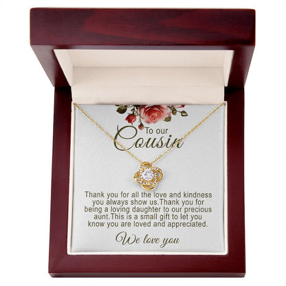 A gold love knot pendant necklace featuring a central round cubic zirconia surrounded by pavé accent crystals. The necklace is presented on a white message card for cousin, inside an open mahogany-finish luxury wooden gift box, accented with red rose artwork at the top.