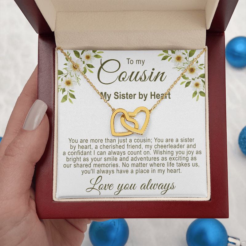 Hand holding an open luxury mahogany-style gift box displaying a gold interlocking hearts necklace with a floral cousin message card, against a background with blue ornaments.