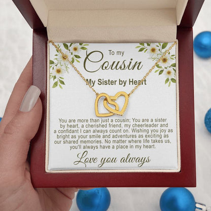 Hand holding an open luxury mahogany-style gift box displaying a gold interlocking hearts necklace with a floral cousin message card, against a background with blue ornaments.