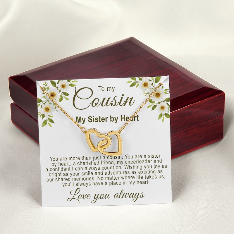 Gold interlocking hearts necklace on a floral cousin message card resting in front of a closed luxury mahogany-style gift box on a white silk background.