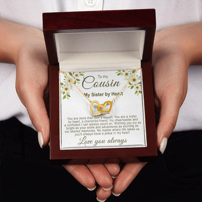 An open luxury mahogany-style gift box held in both hands, presenting a gold interlocking hearts necklace with a floral cousin message card.