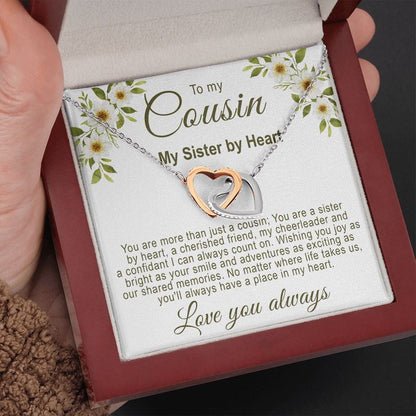 Hand holding an open luxury mahogany-style gift box displaying an interlocking rose gold and silver hearts necklace with a floral cousin message card.