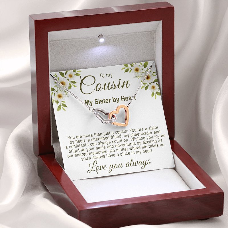 Christmas gifts for your cousin: Illuminated mahogany-style gift box resting on a white silk background, displaying an interlocking rose gold and silver hearts necklace with a floral cousin message card.