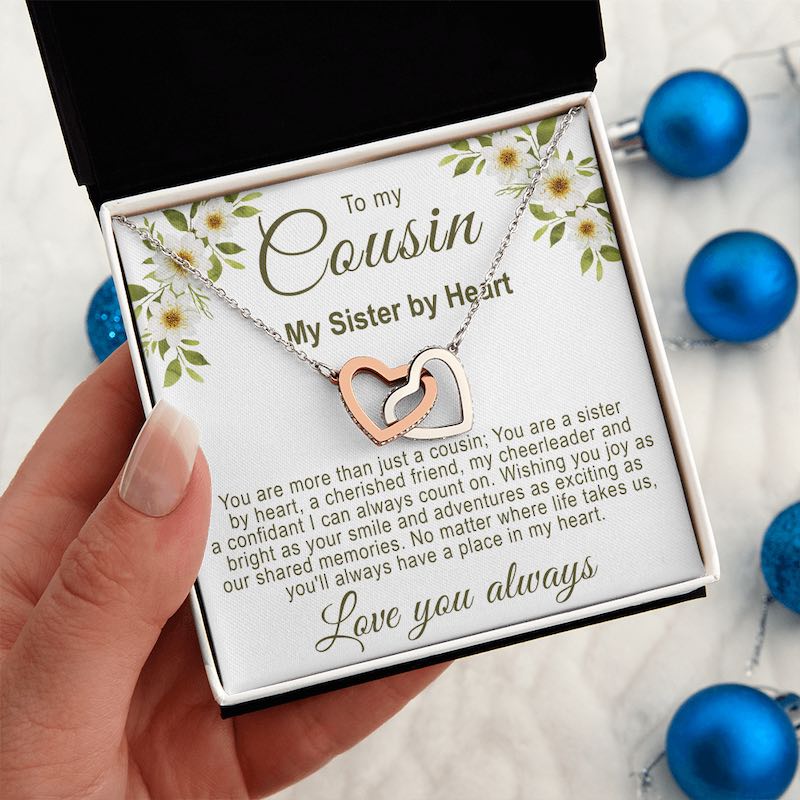 Gift ideas for cousins female: Hand holding a white gift box featuring an interlocking rose gold and silver hearts necklace with a floral cousin message card, against a background with blue ornaments.