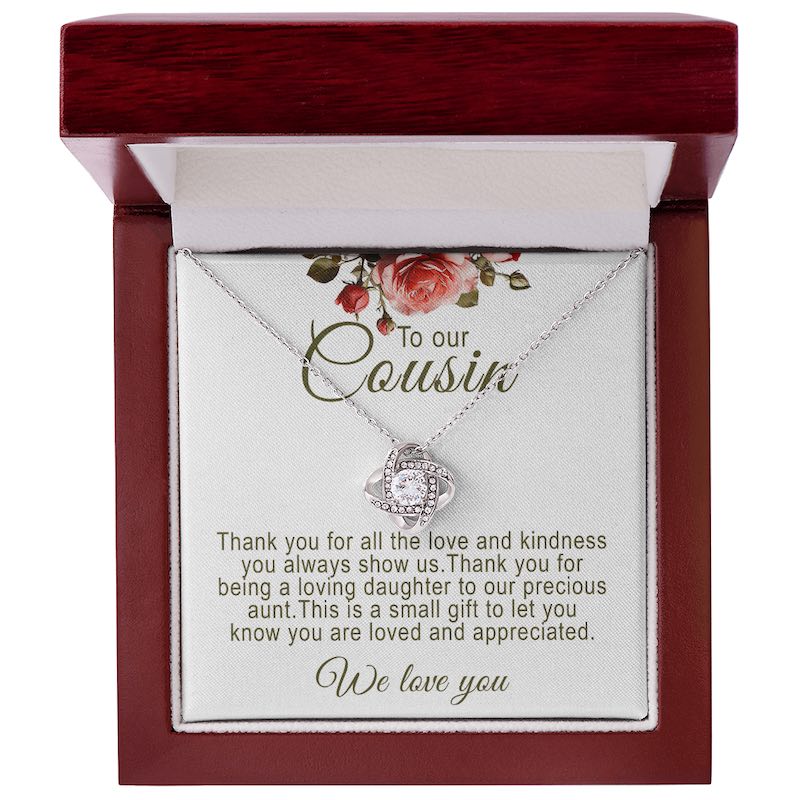 A silver love knot pendant necklace featuring a central round cubic zirconia surrounded by pavé accent crystals. The necklace is presented on a white message card for cousin, inside a mahogany-finish luxury wooden gift box with red rose artwork at the top.