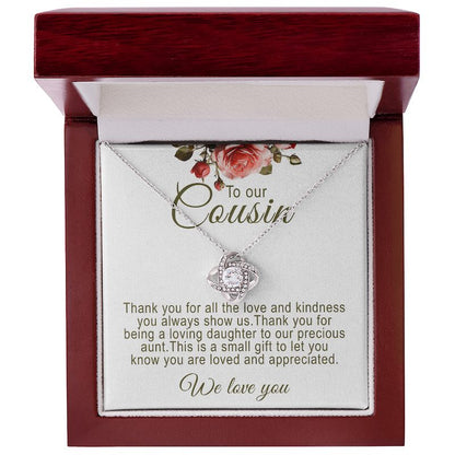 A silver love knot pendant necklace featuring a central round cubic zirconia surrounded by pavé accent crystals. The necklace is presented on a white message card for cousin, inside a mahogany-finish luxury wooden gift box with red rose artwork at the top.