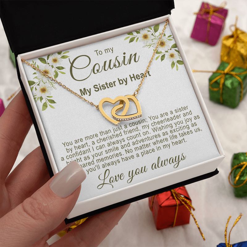 Hand holding a white gift box showcasing a gold interlocking hearts necklace with a floral cousin message card, surrounded by small wrapped gift boxes.