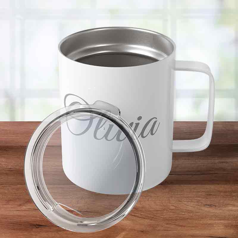 White custom engraved insulated stainless steel coffee mug personalized with "Olivia" in script, sitting on a wooden tabletop with its clear press-in lid leaning against it.