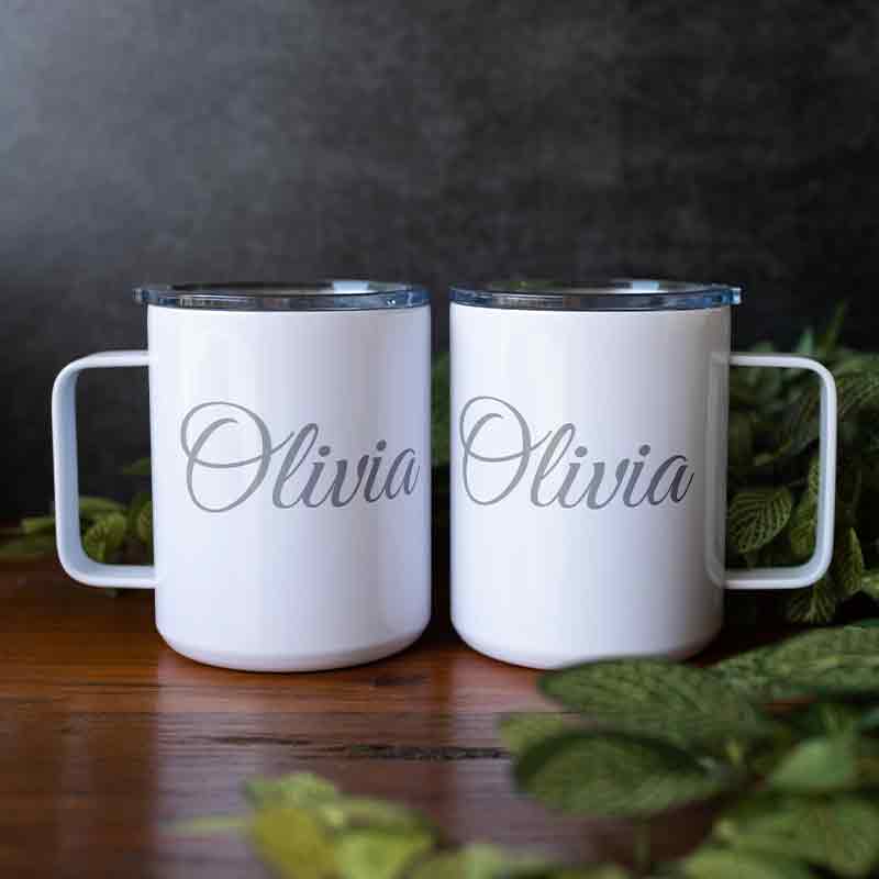 Two white custom engraved insulated stainless steel coffee mugs side by side, both personalized with "Olivia" in script, sitting on a wooden surface with green foliage accents.   