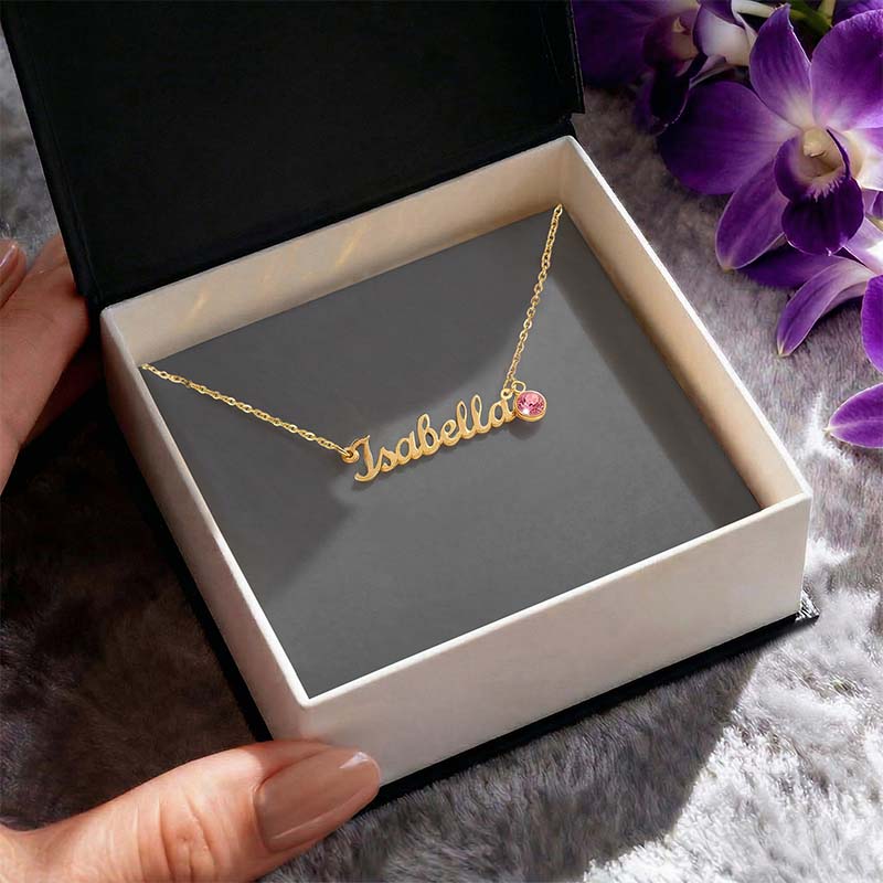 Gold “Isabella” name necklace with a pink gemstone charm displayed inside an open gift box, set on a soft surface with purple flowers in the background