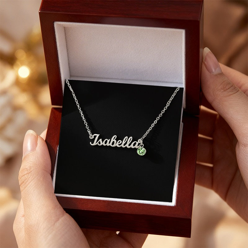 A silver nameplate necklace reading 'Isabella' with a small green gemstone accent, presented in an open dark wood gift box held by two hands.