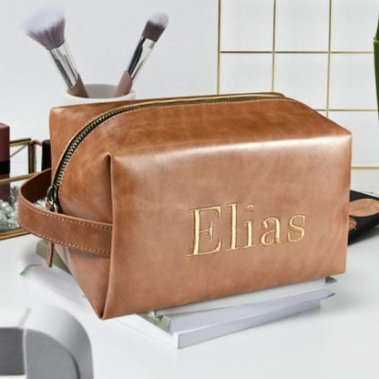 Custom embroidered brown PU leather dopp kit personalized with the name Elias in gold stitching, displayed on a bathroom vanity with cosmetics and makeup brushes.