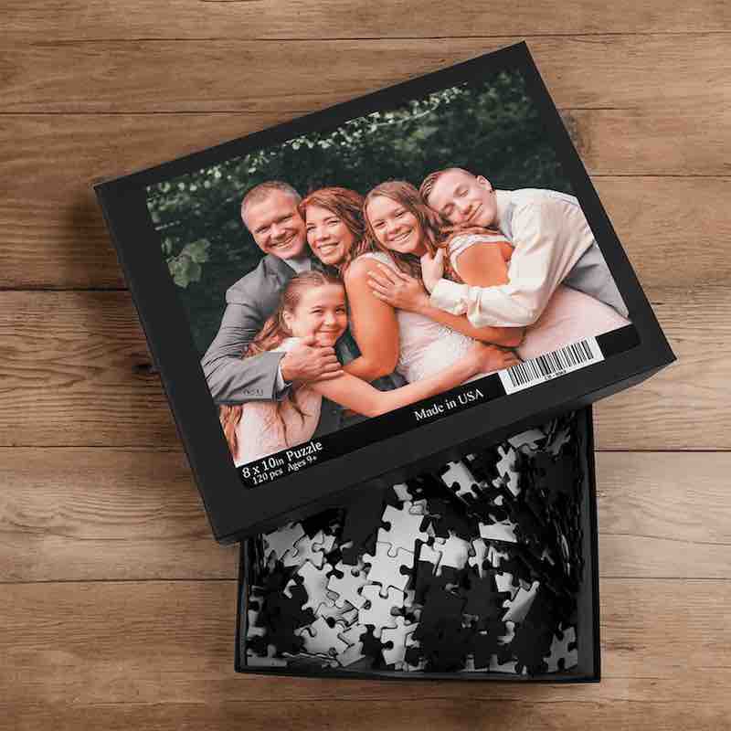 Open 8x10 custom photo puzzle box on a wooden surface filled with loose black-backed puzzle pieces.