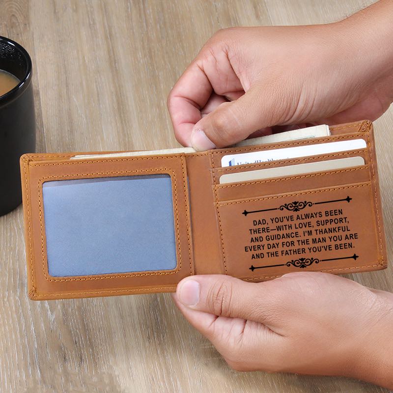 Hands holding an open tan bifold leather wallet on a wooden table, revealing an ID window and card slots engraved with a heartfelt message for Dad.   