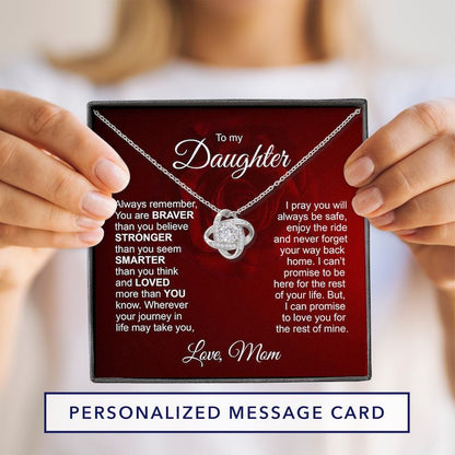 A personalized message card for a daughter, held by hands against a soft blurred background. The card has a dark red background with a silver necklace featuring a heart-shaped pendant with a clear gemstone.