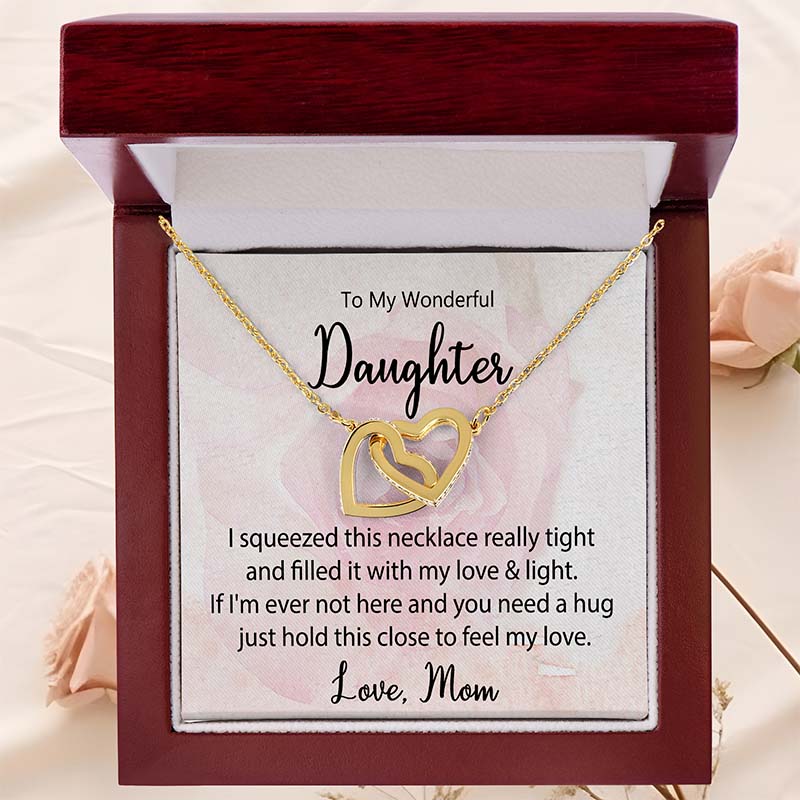 Gold interlocking heart necklace displayed in a luxury mahogany gift box with a heartfelt daughter message card from Mom against a soft blush rose background