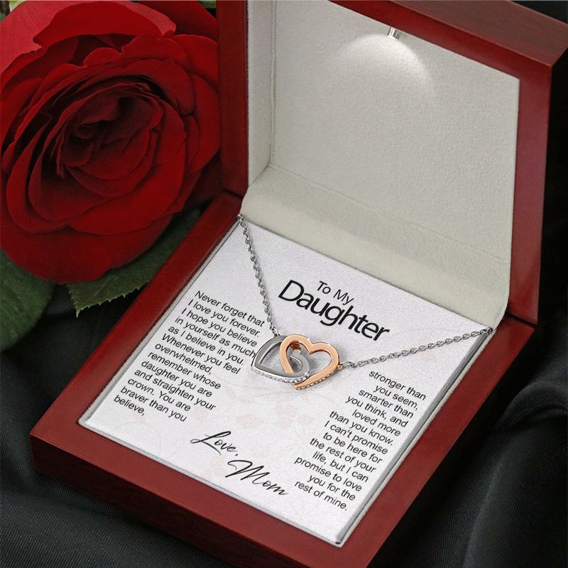 A silver and rose gold necklace featuring two interlocking heart-shaped pendants resting inside an illuminated mahogany-style gift box, set next to a red rose. The necklace is displayed over a white message card titled "To My Daughter" with a sentimental printed message, signed "Love, Mom" in cursive.