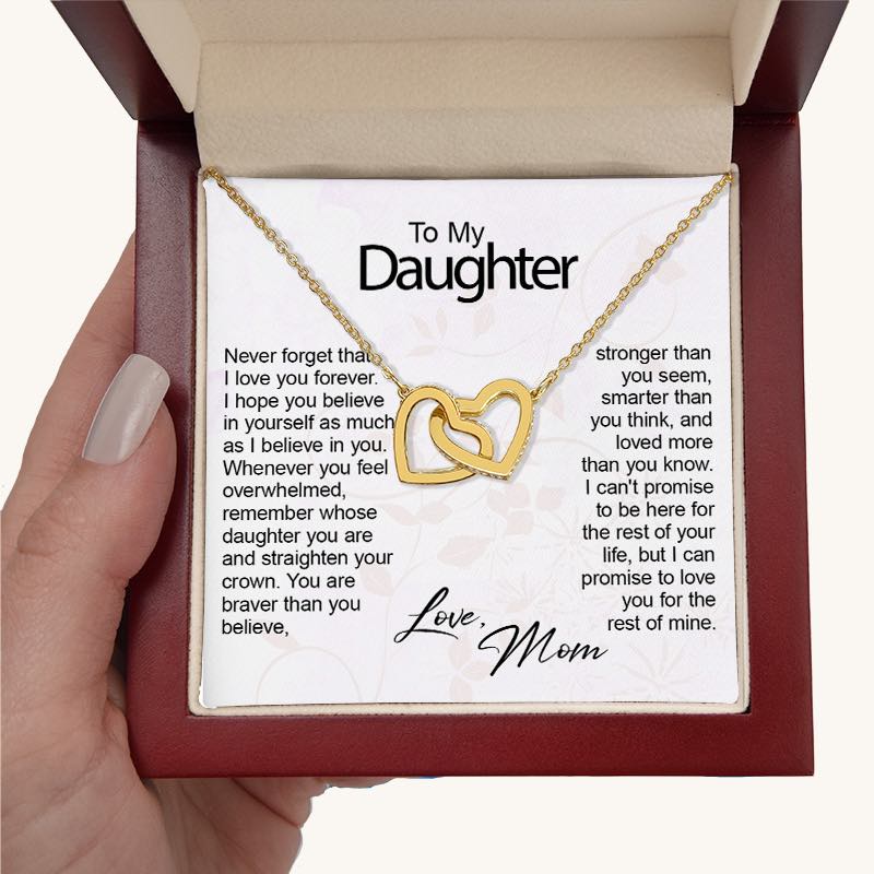 A hand holding an open mahogany-style gift box displaying a gold necklace with two interlocking heart pendants set with small crystals. The necklace rests over a white keepsake card addressed "To My Daughter" with a sentimental printed message and signed "Love, Mom" in cursive script.