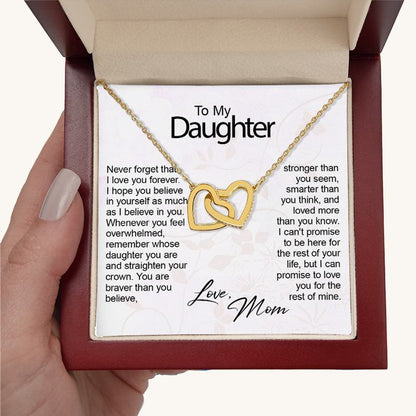 A hand holding an open mahogany-style gift box displaying a gold necklace with two interlocking heart pendants set with small crystals. The necklace rests over a white keepsake card addressed "To My Daughter" with a sentimental printed message and signed "Love, Mom" in cursive script.