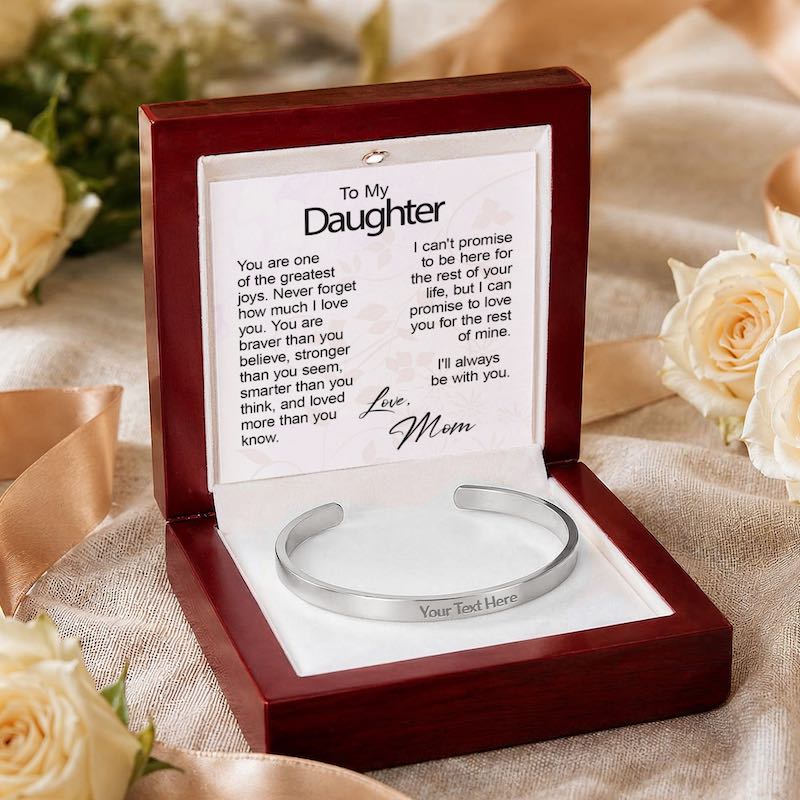 Personalized silver cuff bracelet with "Your Text Here" sample engraving, presented in an open wooden gift box resting on textured beige silk beside white roses, featuring a "To My Daughter" card from Mom.   