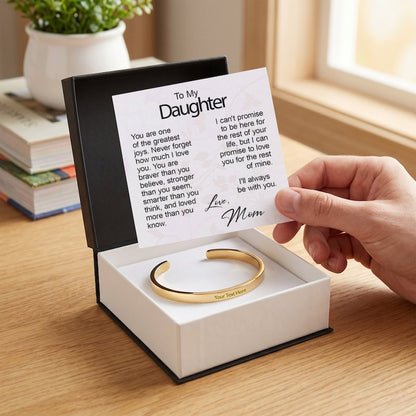 A hand holding open a printed "To My Daughter" message card inside a gift box featuring a custom-engraved silver cuff bracelet, set on a wooden table near a window.